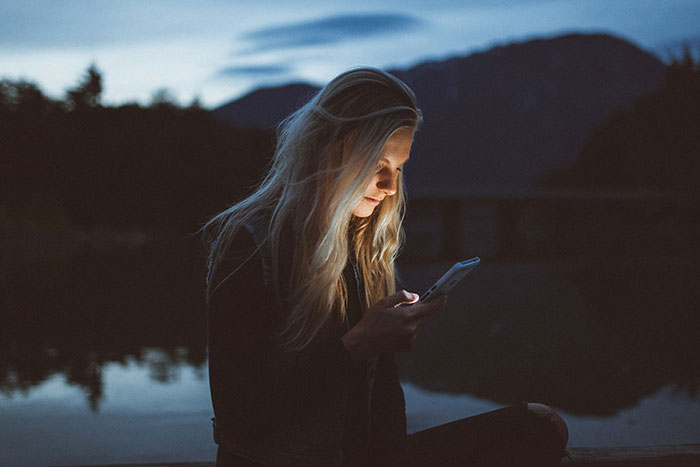 A woman sitting by a lake at dusk, looking at her phone, creating a mysterious atmosphere.