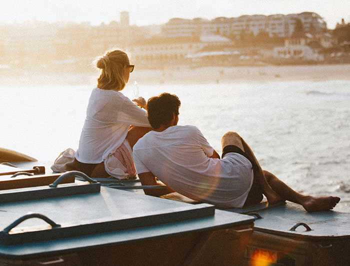 Couple sitting next to each other on a rooftop overlooking the ocean during sunset, wearing casual white clothing.