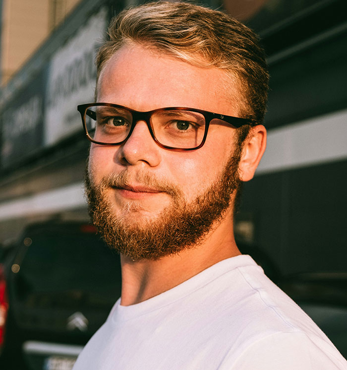 Man with glasses and a beard wearing a white shirt, seated outdoors, capturing a casual moment with neutral expression.