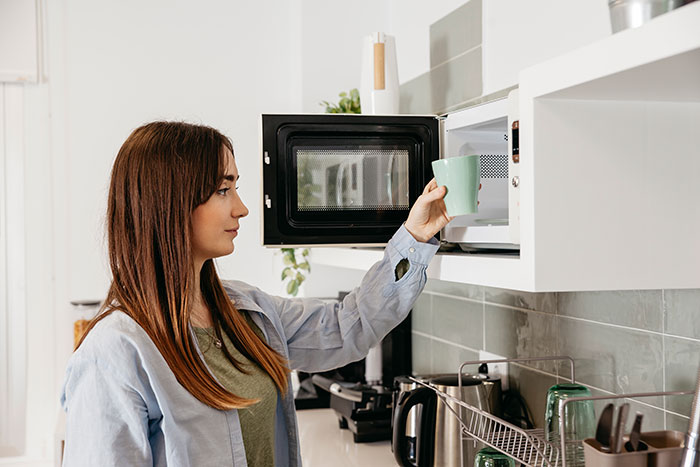 Woman using a microwave in the kitchen, wearing a light blue shirt, holding a green mug.