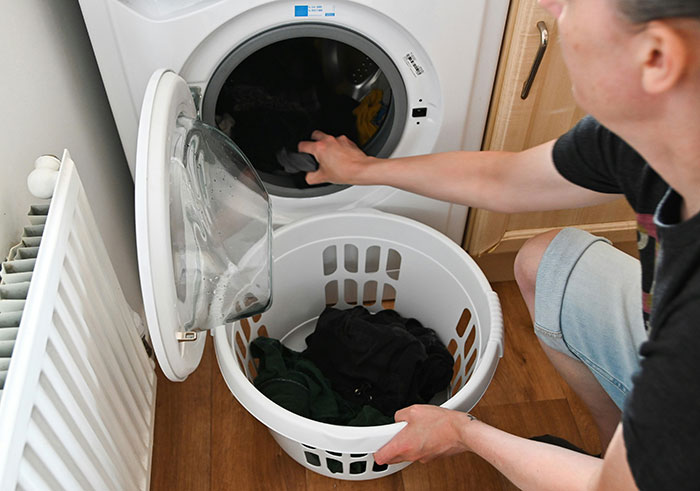 Person loading clothes into a washing machine, holding a laundry basket on the floor.