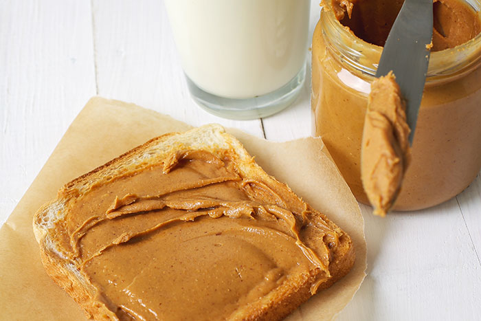 Peanut butter spread on bread with a knife, next to a jar and a glass of milk on a white background.