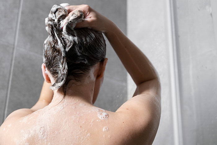 Person washing hair with shampoo in the shower, creating lather while cleansing scalp.