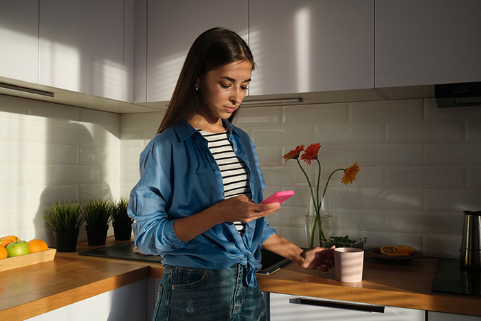 Young woman in kitchen using smartphone, holding a pink mug, with sunlight on her face.