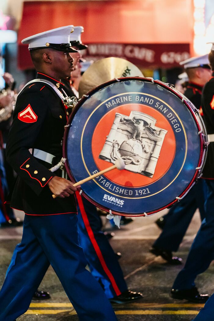 Marine Band San Diego member marches, showcasing uniform and large drum in a parade.