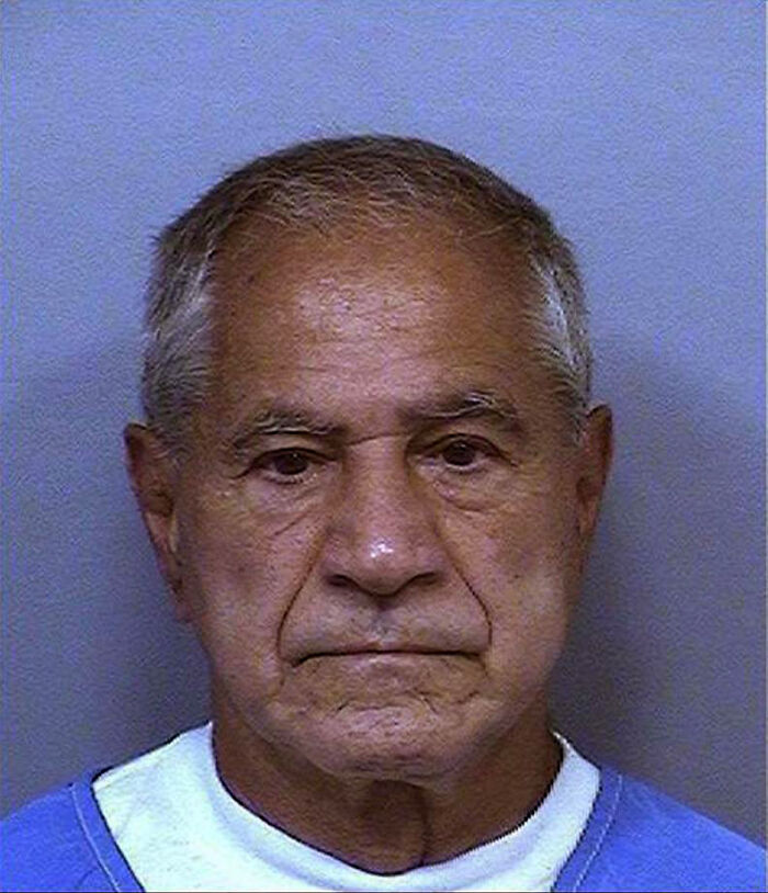 An older man with a serious expression wearing a blue shirt, photographed against a plain backdrop.