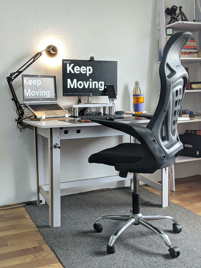 Desk setup with screens displaying "Keep Moving," a black chair, and books on a shelf.