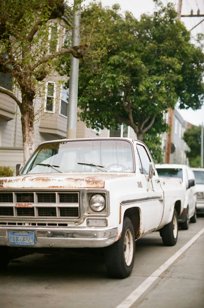 Rusty GMC truck parked on a city street, surrounded by trees and buildings.