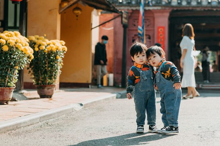 Two young children in denim overalls standing on a street, surrounded by yellow flowers, capturing a heartwarming moment.