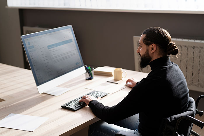 Man with a beard working at a desk with a computer, sipping coffee in an office setting, focused on work tasks.