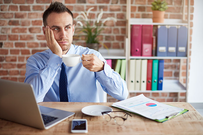 Man in an office looking thoughtful with coffee, open laptop, and documents on desk.