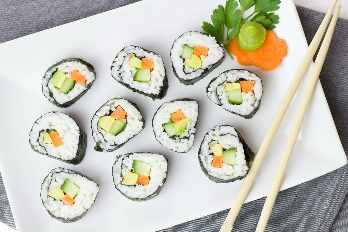 Sushi rolls with carrot and cucumber on a white plate, accompanied by garnish and chopsticks, highlighting unhealthy food.