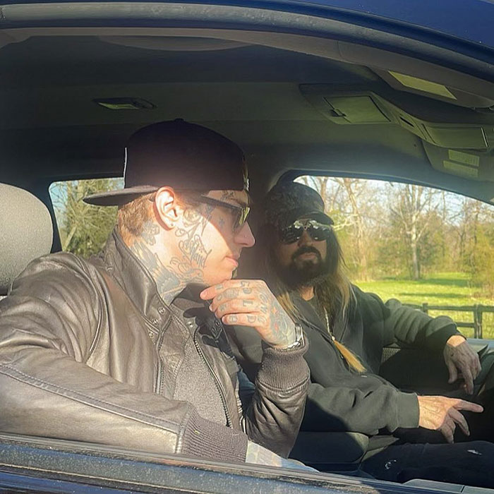 Two men sitting in a parked car, one with tattoos and a hat, the other with a beard and sunglasses, in a sunny setting.