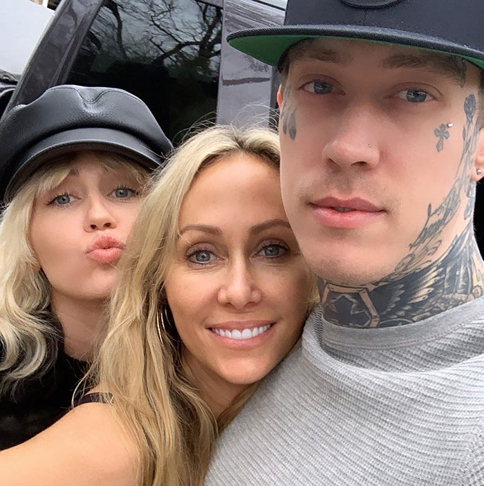 A trio posing for a selfie outdoors, featuring Miley Cyrus' brother with tattoos and wearing a cap.