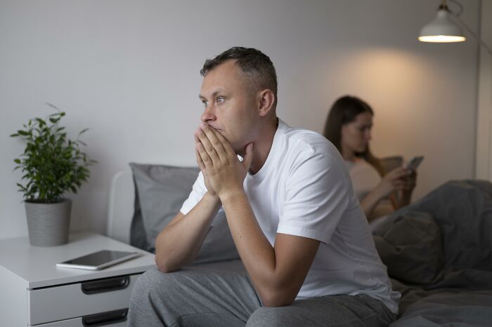 Man in deep thought sitting on bed, woman using phone in the background, reflecting an unreasonable friend relationship.