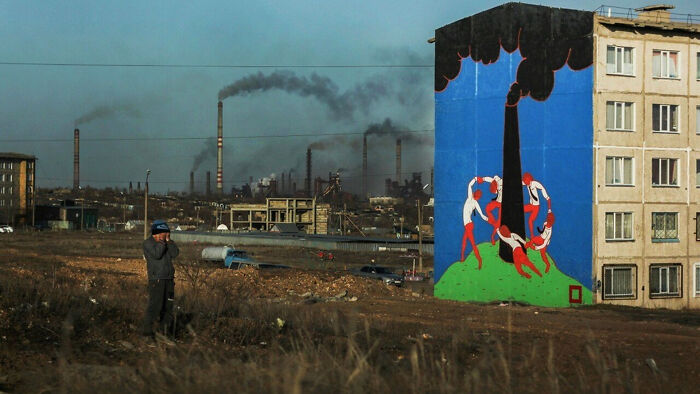 Urban hell scene with industrial smoke and a colorful mural on a building, depicting contrast amidst pollution.