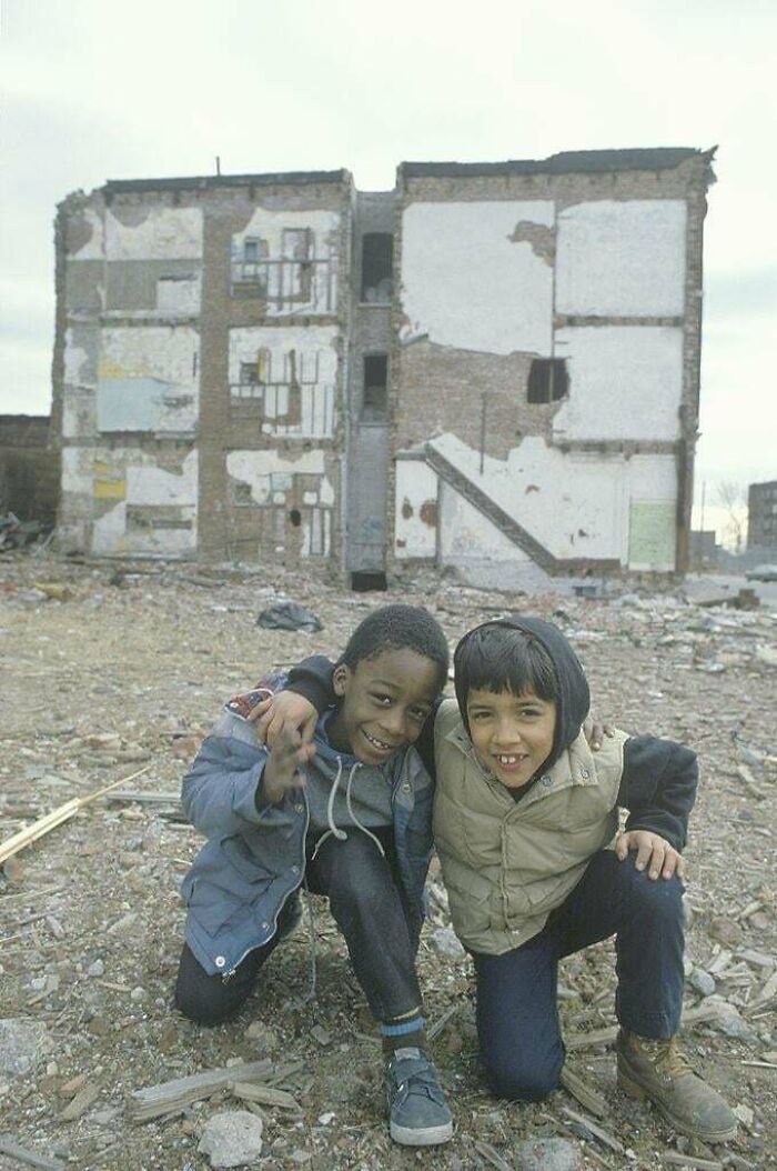 Two children smiling in front of a deteriorating apartment building in an urban hell environment.