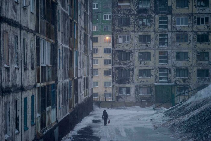 A lone figure walks between decaying apartment buildings in a bleak urban hell setting.