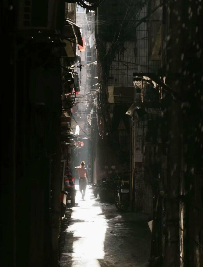 A person walking through a narrow, dark urban alley illuminated by sunlight.