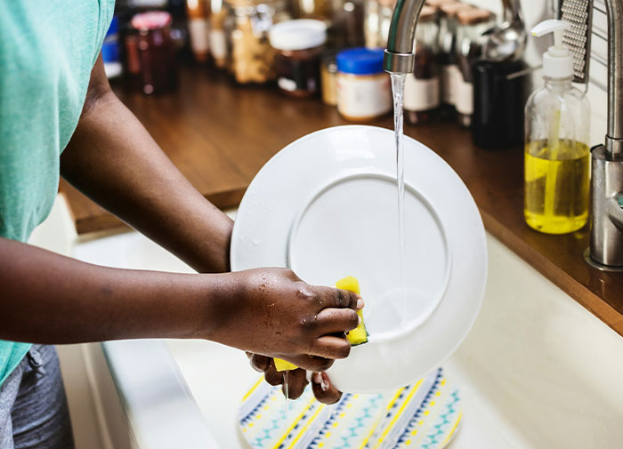 Person washing a plate with running water, showcasing using things the wrong way in kitchen cleaning.