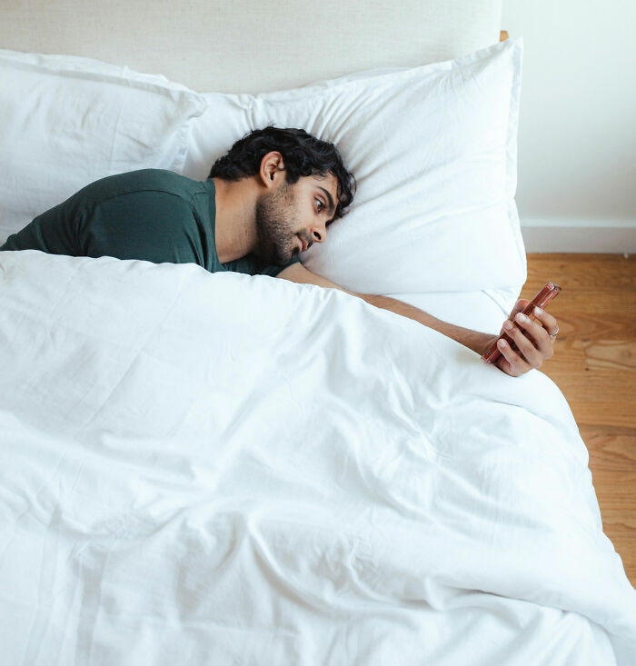 Man lying in bed, casually holding a smartphone, exhibiting non-sexual behavior that attracts women.