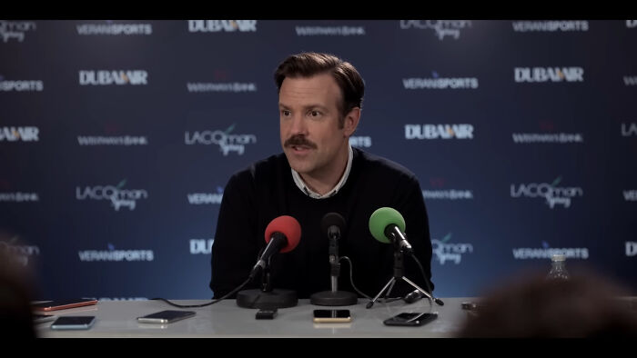 Man at a press conference table, speaking into microphones, with a blue backdrop showing various sponsor logos.