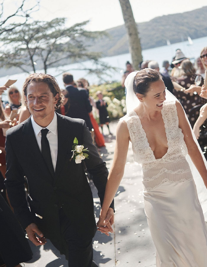 Australian bride in a wedding dress, holding hands with groom, surrounded by guests outdoors.