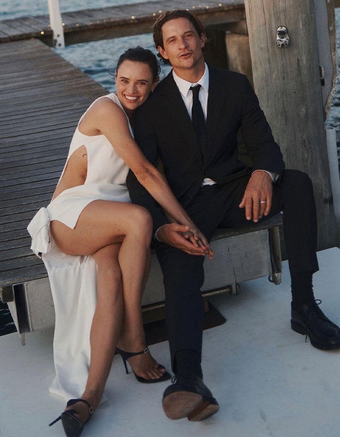 Australian bride in white dress sitting by a dock with groom, holding hands and smiling.