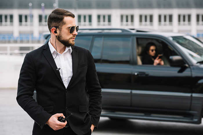 Bodyguard in a suit with earpiece, standing near a luxury car with a passenger using a phone, showcasing work for the wealthy.