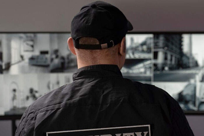 Back view of a bodyguard monitoring security cameras, wearing a black cap and jacket in a control room setting.