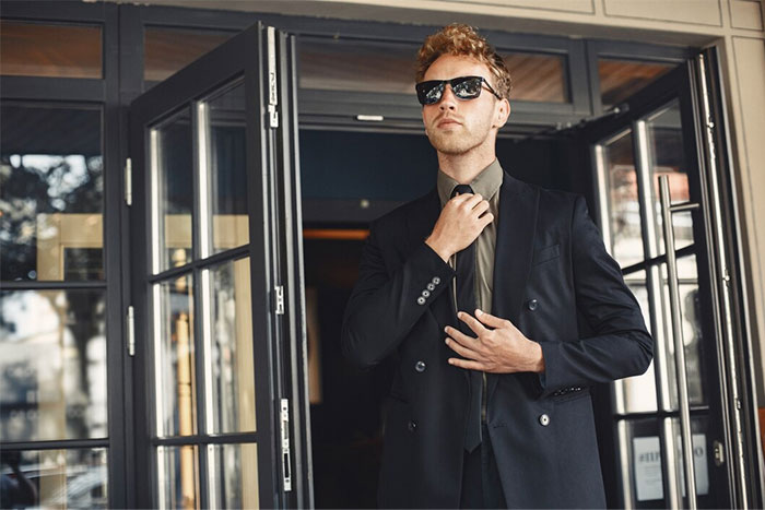 Man in a suit and sunglasses adjusting his tie, embodying the style of bodyguards for the wealthy.