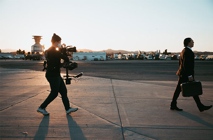 A bodyguard in a suit carrying a briefcase walks beside a filmmaker at sunset on an airport runway.