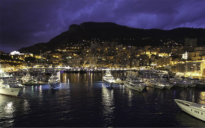 Monaco harbor at night, luxury yachts docked, city lights illuminating the wealthy lifestyle.