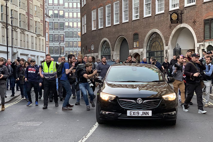 A bodyguard escorts a vehicle through a crowd of photographers on a busy city street.