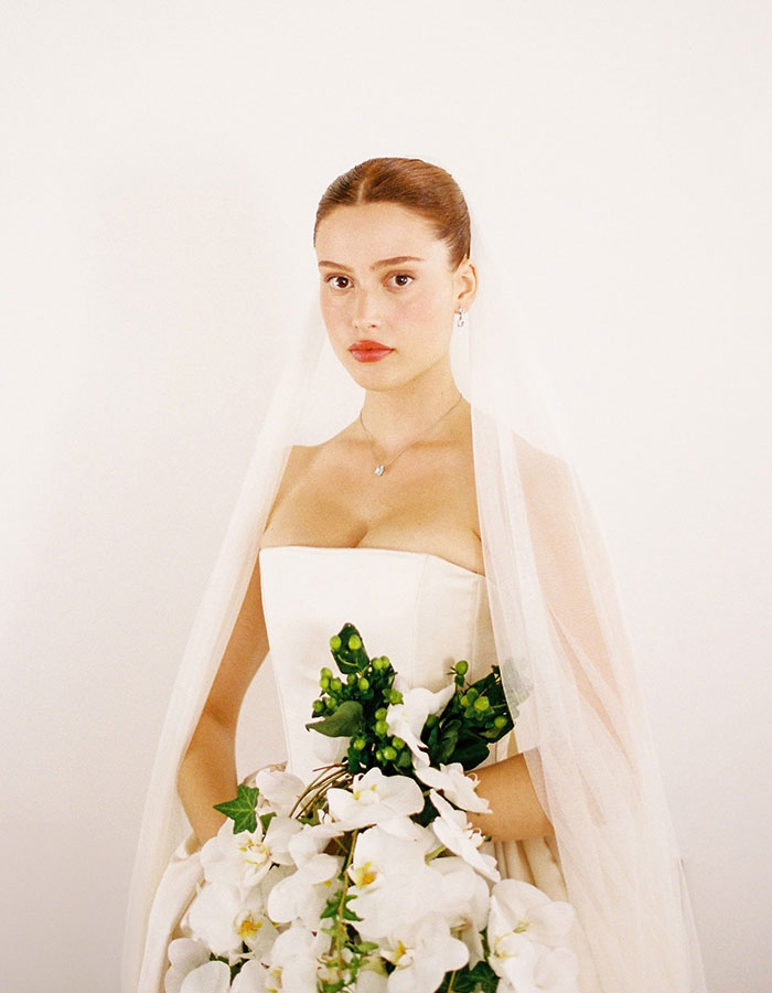 Woman in dramatic wedding dress and veil, holding white flower bouquet, standing against a plain background.