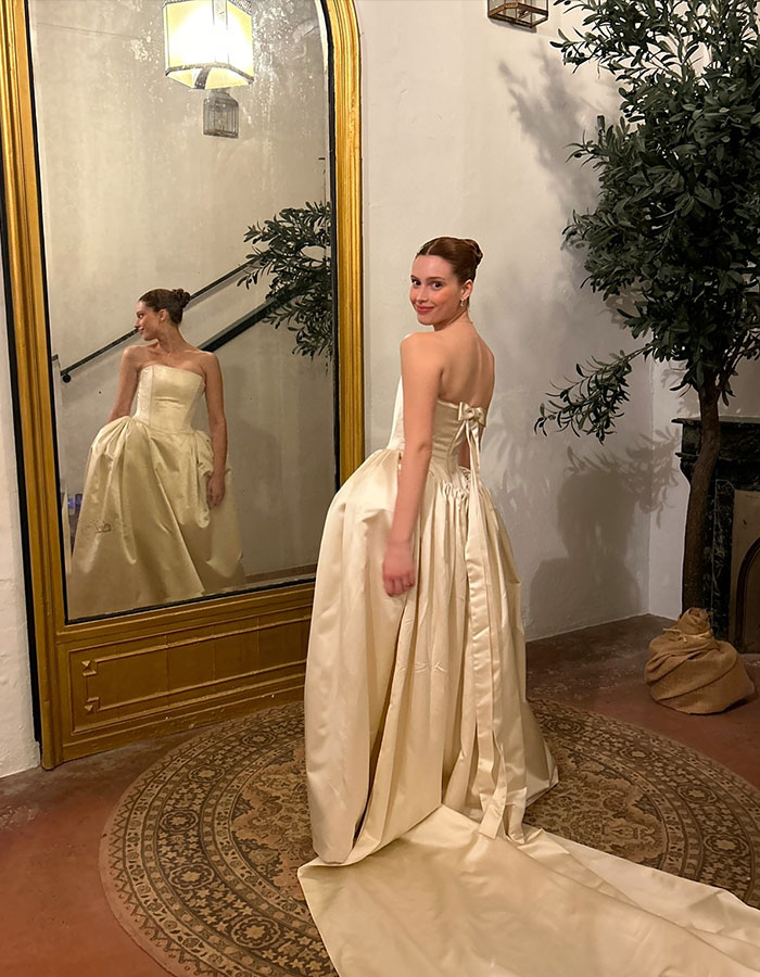 Woman in dramatic wedding dress with veil, standing by a large mirror, displaying the back of the gown in a stylish setting.