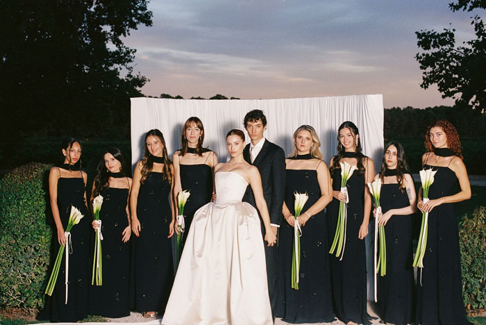 Bridal party posing, bride in a dramatic wedding dress and veil, with bridesmaids in black dresses holding white flowers.