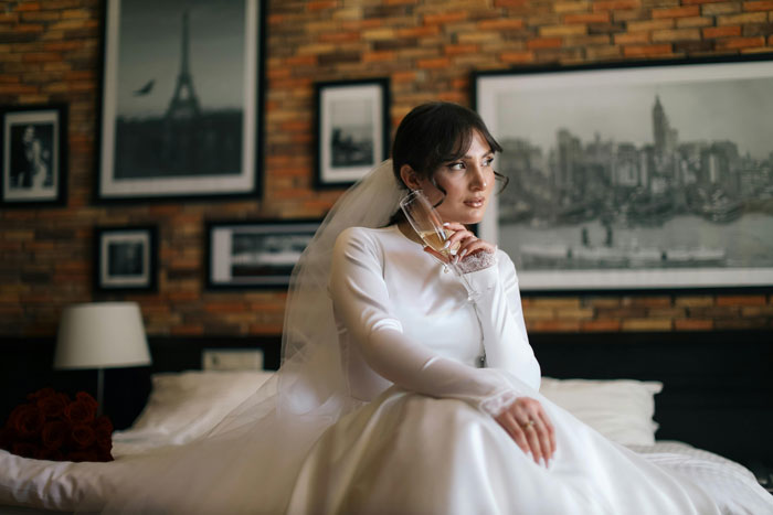 Bride in a white dress and veil, sitting on a bed, holding a champagne glass, in a room with framed pictures on the wall.