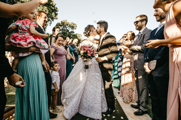 Wedding celebration with bride and groom kissing, surrounded by guests in colorful attire, outdoors on a sunny day.