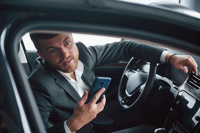 Man in a suit talking on the phone inside a car, possibly discussing rich people stories while driving.