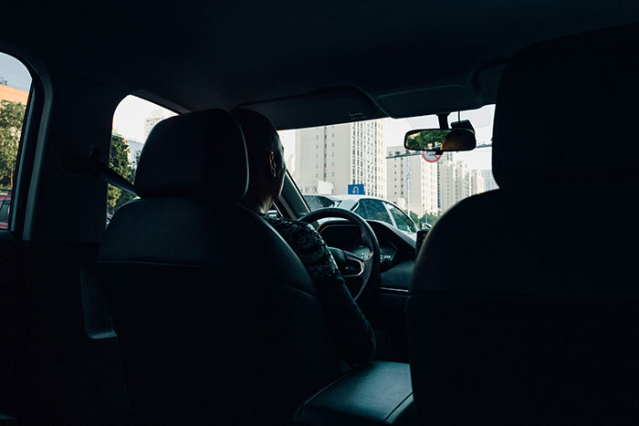 Driver in a city car, capturing stories about wealthy individuals, with urban buildings visible through the windows.