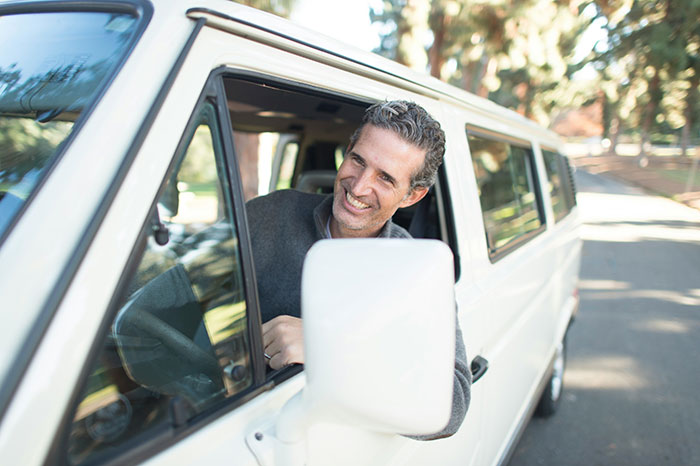 A smiling driver in a white van, possibly hearing stories about wealthy passengers while on duty.