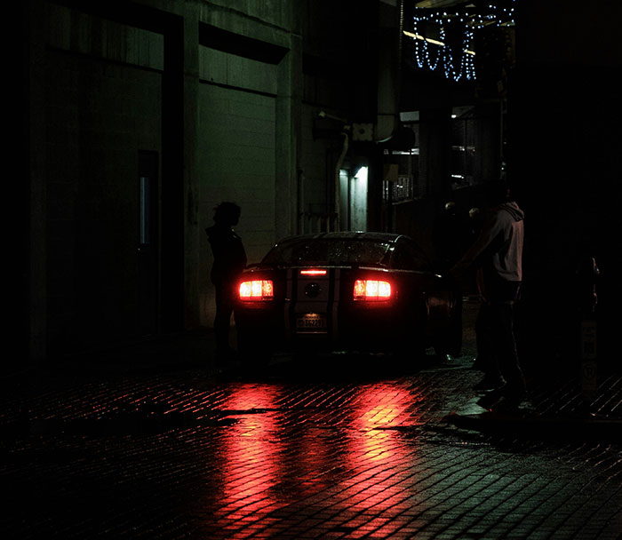 Car in a dimly lit alley with reflections on wet pavement, capturing a mysterious ambiance about driving rich people.