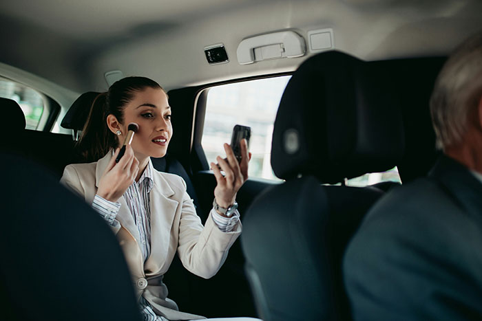 Woman applying makeup in car, seated behind the driver, embodying stories from drivers about rich people.