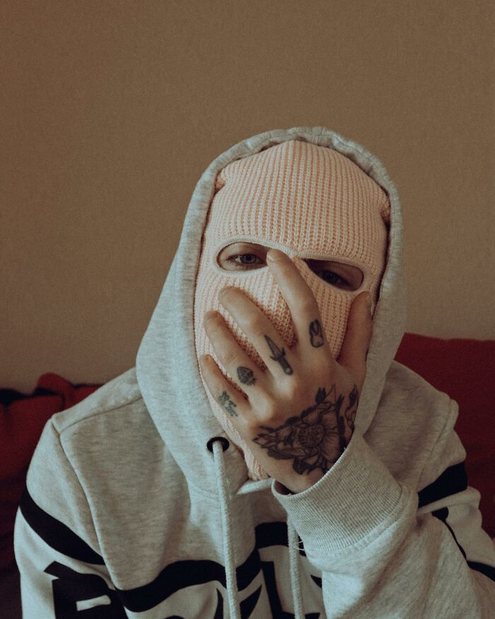 Person in a knit mask with tattooed hand over face, wearing a hoodie, capturing an awkward delivery moment.