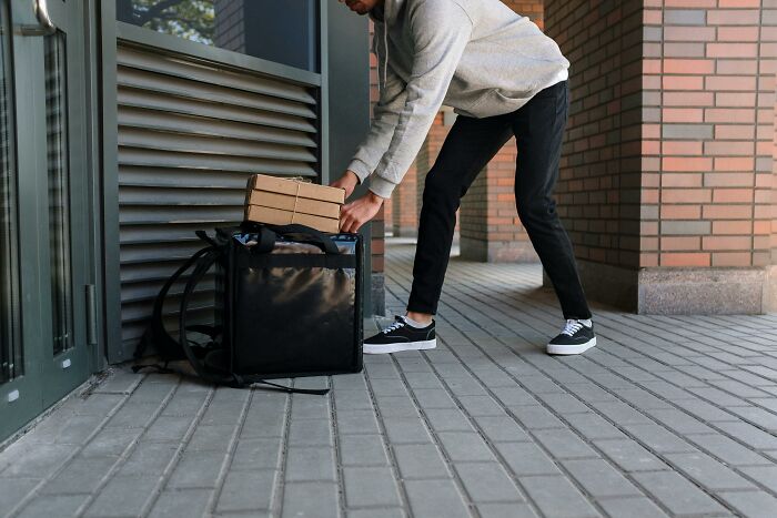 Delivery person in a hoodie crouching to place boxes on the ground beside a brick building.