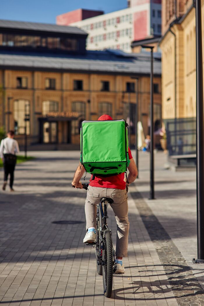 Delivery cyclist with a large green backpack riding through a city street in daylight.