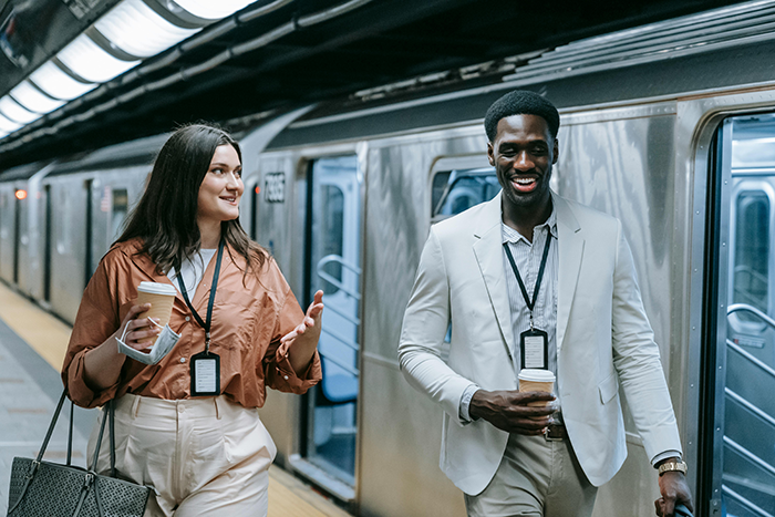 A man and woman in business attire walking in a subway station, holding coffee and talking, both wearing ID badges.