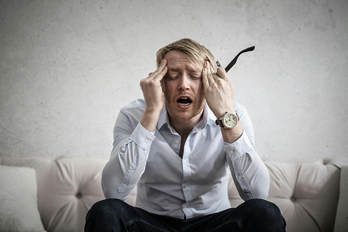 Man in distress on a sofa, wearing a white shirt and holding his head, embodying issues with keeping privacy.