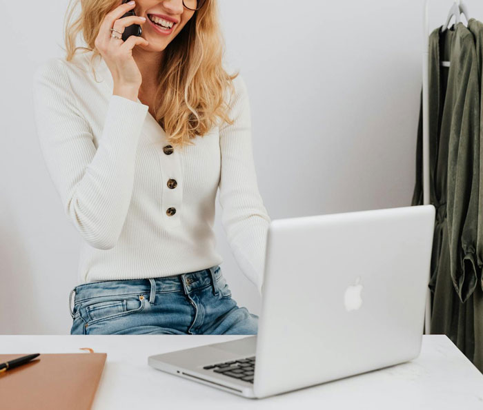 Woman smiling, talking on phone, using laptop at desk, related to revenge theme.